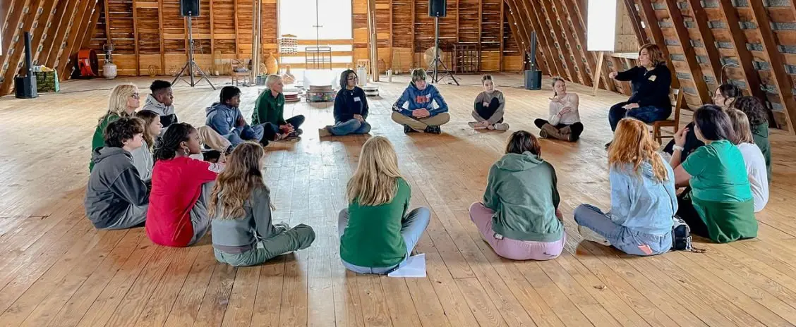 Group of 15-18 teens and adults sit in a circle on a barn floor listening to a woman speaking
